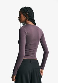 Long-sleeve purple top in soft fabric with a fitted design. Features a round neckline and subtle seam detailing, paired with dark pants.
