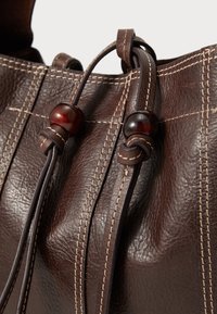Sac en cuir marron avec surface texturée, coutures claires contrastantes et accent en perles en bois sur les cordons de serrage. Design minimaliste.