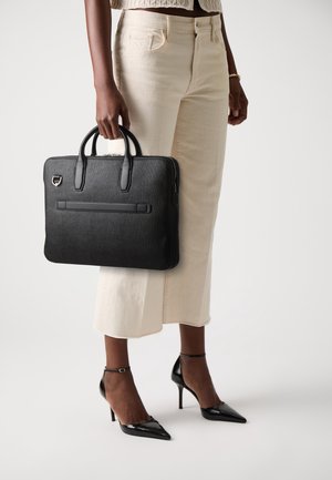 Personne tenant une mallette en cuir noir texturée, portant un jean large crème, des escarpins noirs à bout pointu et un pull court beige.