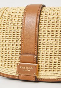 Kudottu olkakotelo, jossa on vaaleanruskea nahkahihna ja metallilevy, johon on painettu "kate spade NEW YORK". Ominaisuuksia ovat sileä pinta ja siisti ommel.