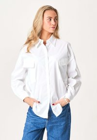 Kvinde med langt blondt hår iført en knaplukket hvid skjorte og blå jeans, med hænderne i lommerne, ser til siden mod en ensfarvet baggrund.