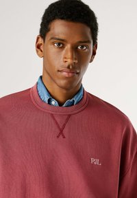 Sudadera burdeos con escote acanalado y logo de PJL bordado. Superpuesta sobre una camisa de denim azul claro. Tejido suave y texturizado.