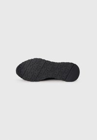 Semelle de chaussure noire avec motif répété sur la bande de roulement et nom de marque embossé près du talon, présentée sur un fond clair uni.
