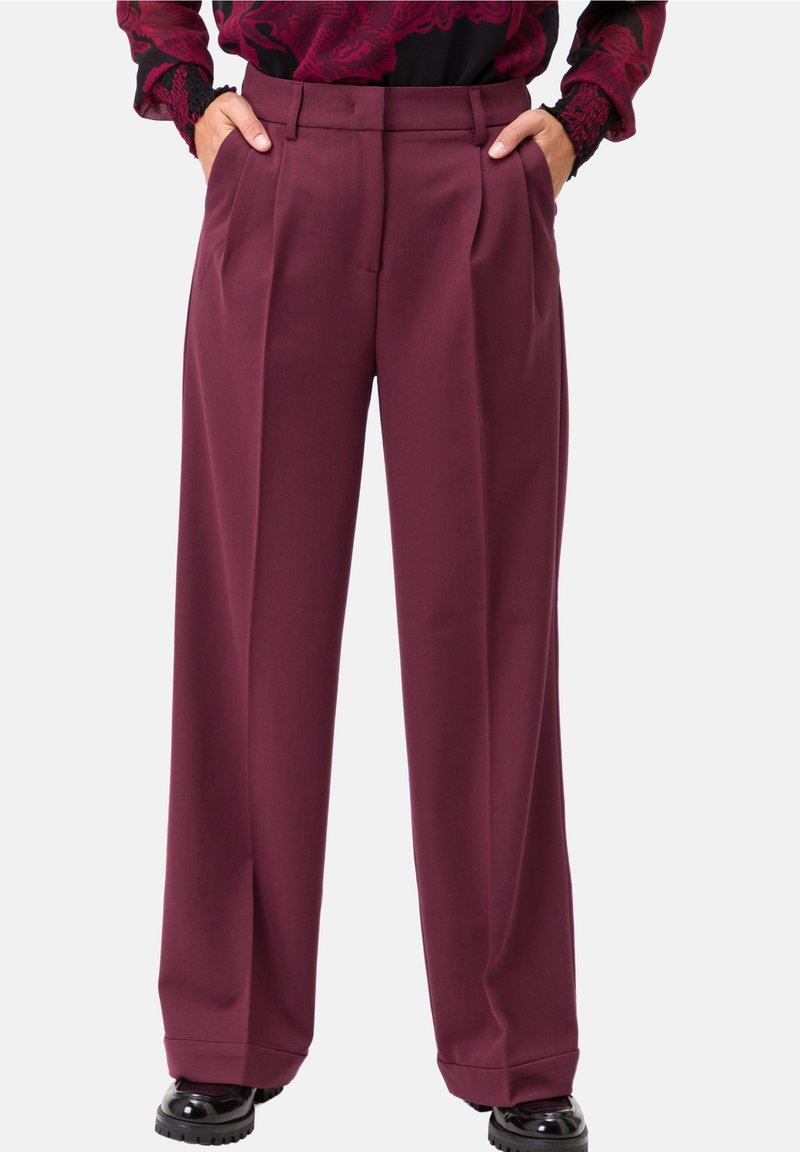 Personne portant un pantalon bordeaux taille haute à jambes larges, mains dans les poches, et des chaussures noires brillantes, visible de la taille aux pieds.