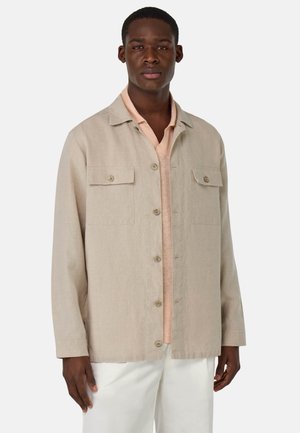 Giacca camicia di lino beige con una vestibilità rilassata, caratterizzata da due tasche sul petto, chiusura a bottoni e un tessuto morbido e strutturato, indossata sopra un top pesca.