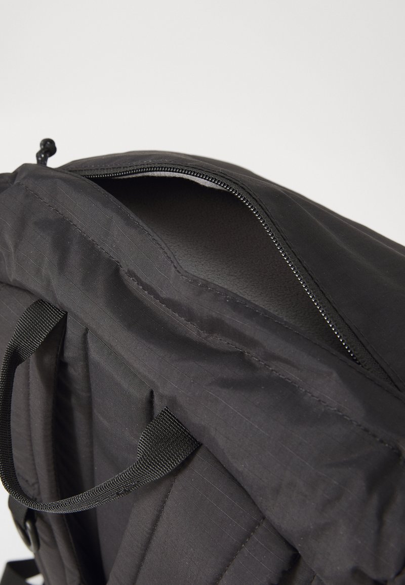 Sac à dos noir au texture lisse, comportant une ouverture zippée, des bretelles plates et un design en panneaux. Le matériau semble durable et résistant à l'eau.