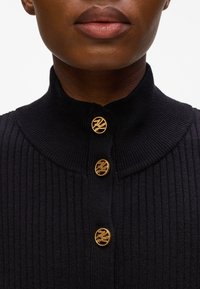 Top in maglia nera a coste con colletto alto e tre bottoni decorativi dorati con design del logo. Tessuto strutturato e stile aderente.