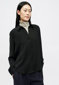 Žena s dugom tamnom kosom nosi crnu košulju s dugim rukavima preko prugastog rolka, stoji s rukama u džepovima na bijeloj pozadini.