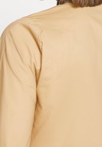 Giacca leggera beige con una texture liscia, caratterizzata da un design semplice e dettagli minimi. Il tessuto appare morbido e leggermente lucido.