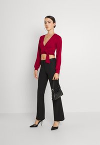Top cropped de mangas compridas vermelho com nervuras e nó frontal, combinado com calças pretas à boca de sino e saltos pretos de bico fino, segurando uma pequena bolsa preta.