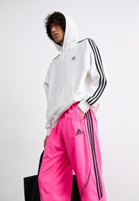 Hoodie branco com design de três riscas pretas, adornado com um pequeno logotipo. Calças rosas brilhantes com detalhes e logotipo pretos. Textura de tecido suave.