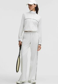 Valge sportlik tracksuit, mis on valmistatud kergetest kangast, mustade aksentriidega, kõrge kraega ja mugava lõikega pükstega. Hoides tennisereketit.