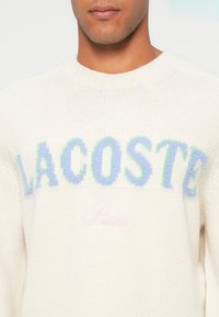 Bărbat îmbrăcat cu un pulover alb tricotat pe care scrie "LACOSTE" cu litere pufoase albastre și verzi pe piept.