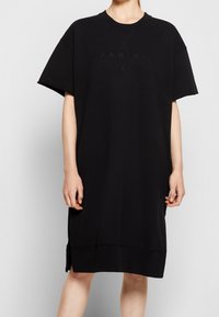 Vestido t-shirt oversized negro, confeccionado en algodón, con mangas cortas. Presenta un logotipo de la marca en relieve y aberturas laterales en el dobladillo.