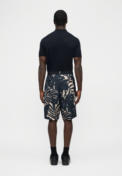 Άνδρας που στέκεται με την πλάτη στραμμένη, φοράει μαύρο κοντομάνικο πουκάμισο και ναυτικού χρώματος σορτς με μεγάλα μπεζ φύλλα και μαύρα παπούτσια.