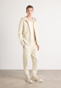 Jonge man die staat te poseren in een beige hoodie, bijpassende joggingsbroek, wit geribbeld tanktop en beige sneakers tegen een effen achtergrond.