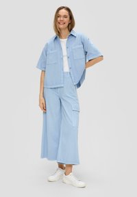 Helles blaues Jeanshemd mit kurzen Ärmeln und zwei vorderen Taschen, kombiniert mit weiten hellblauen Hosen und weißen Turnschuhen.