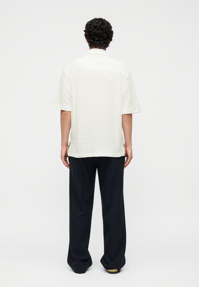 Άνδρας με σγουρά μαλλιά στέκεται με την πλάτη, φοράει λευκό, υφασμάτινο, κοντομάνικο πουκάμισο και χαλαρό μαύρο παντελόνι σε μονόχρωμο φόντο.