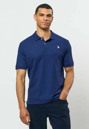 Homme portant un polo bleu marine à manches courtes avec un petit logo blanc brodé sur la poitrine gauche, debout avec une main dans la poche.