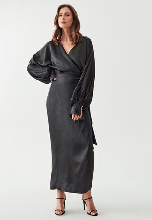 Black wrap dress featuring a textured, satin-like fabric, long balloon sleeves, and a fitted silhouette. Accompanied by black heeled sandals.