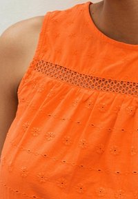 Top de algodón naranja con flores bordadas y detalles de corte geométrico a lo largo del corpiño. La tela es suave con un acabado ligeramente texturizado.