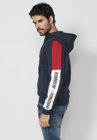 Jūrininkų sweatšertas su raudonais ir baltais juostuotais rankovėmis bei logotipu, su užtraukiama kišene ir reguliuojamu kapučinu. Minkšta, tekstūruota medžiaga.