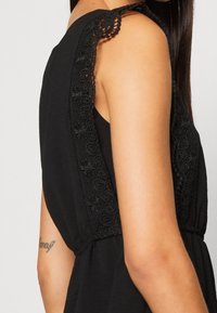 Vestido negro con adorno decorativo de encaje en el hombro, textura suave y cintura entallada. Vista lateral que muestra el diseño y los detalles.