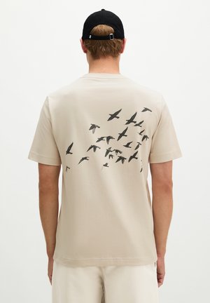 Mann trägt beige T-Shirt mit schwarzem fliegendem Vogeldruck auf dem Rücken und einer schwarzen Baseballkappe, Rücken zur Kamera.