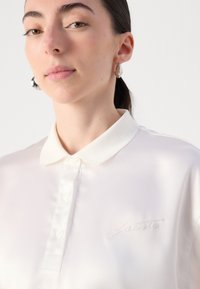 Polo blanc avec une finition satinée, doté d'une patte de boutonnage et d'un logo brodé subtil sur la poitrine gauche. Texture lisse et coupe ajustée.