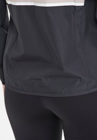 Svart vindjacka med matt finish, med ett vitt och beige rand högst upp och "ENDURANCE"-logotypen på nederdelen av ryggen.