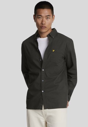 Camicia button-up verde scuro in cotone, con tasche laterali e un piccolo logo giallo sulla tasca del petto, indossata sopra una maglietta bianca.