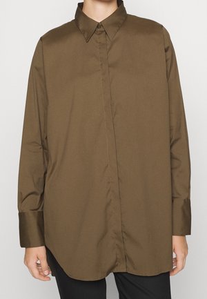 Løst siddende langærmet olivengrøn skjorte med klassisk krave, båret over sorte bukser, der viser mave og arme.