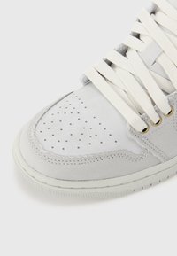 Gros plan sur le bout d'une basket blanche et gris clair avec cuir perforé, bordure en tissu, lacets crème et œillets dorés sur fond blanc.