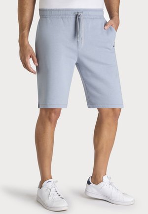 Uomo che indossa pantaloncini azzurri con coulisse e sneakers Adidas bianche, in piedi davanti a uno sfondo semplice.