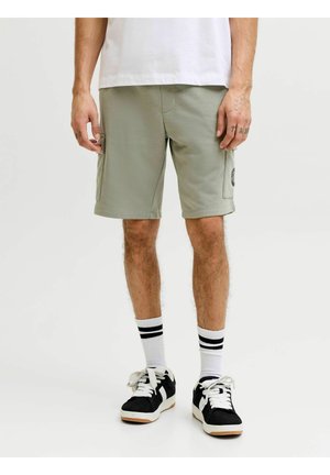 Uomo che indossa pantaloncini cargo verde chiaro, calzini bianchi con strisce nere, scarpe da ginnastica nere e bianche, in piedi davanti a uno sfondo neutro.