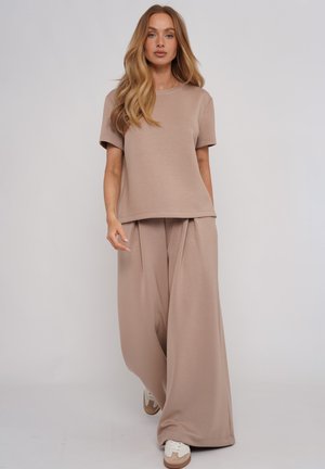 Kobieta o długich falujących włosach, ubrana w luźny beżowy krótki top i szerokie spodnie, idzie do przodu w białych trampkach na jednolitym tle.