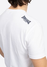 Hvid bomulds t-shirt med et blåt broderet logo på den øverste del af ryggen. Korte ærmer og et klassisk rundt halsdesign.