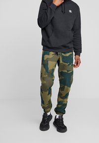 Black hoodie with adjustable drawstring, paired with green and blue camo joggers, featuring an elastic waistband and cuffs, worn with black sneakers.