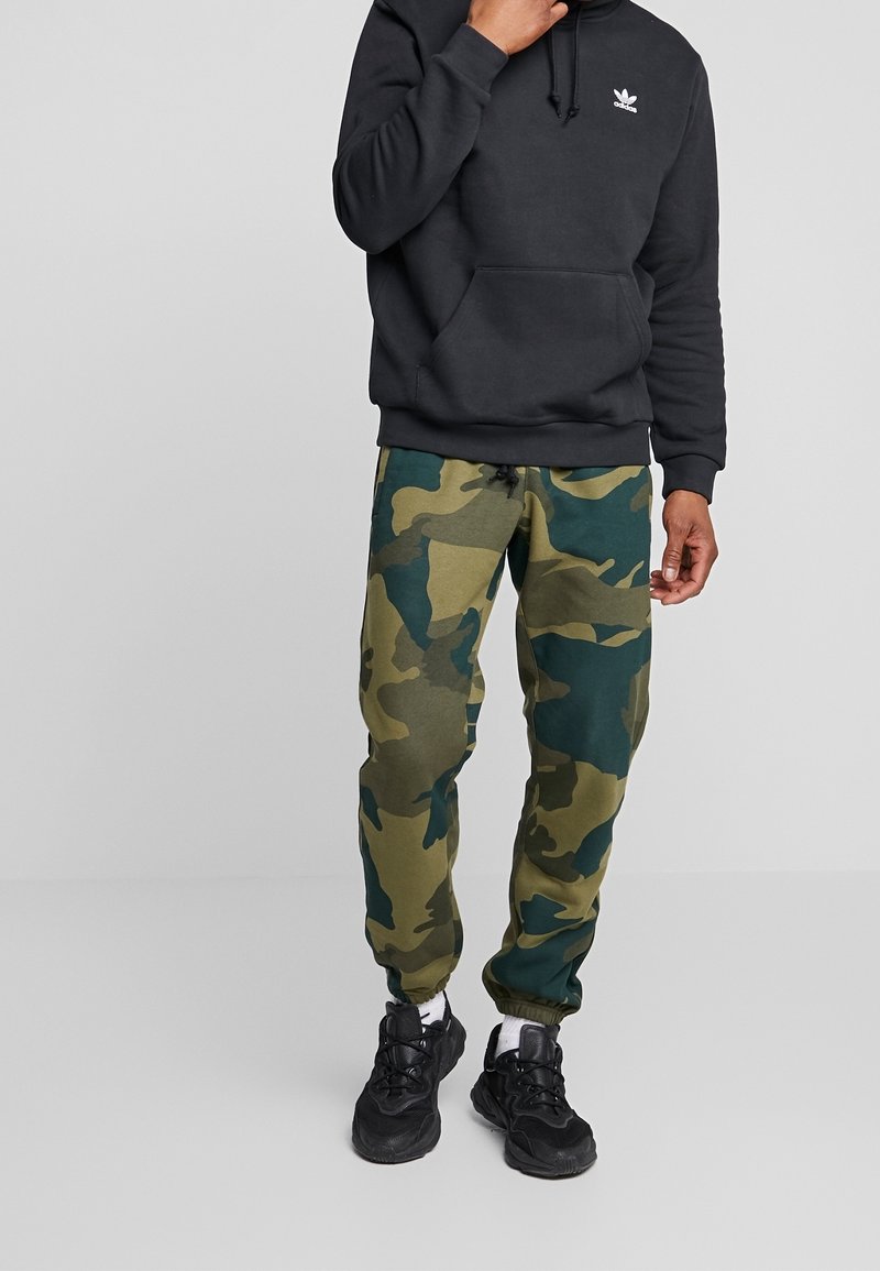 Black hoodie with adjustable drawstring, paired with green and blue camo joggers, featuring an elastic waistband and cuffs, worn with black sneakers.