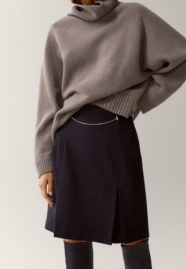 Personne portant un pull à col roulé gris rentré dans une jupe noire longueur genou avec une fine ceinture en chaîne et des bottes noires montantes.