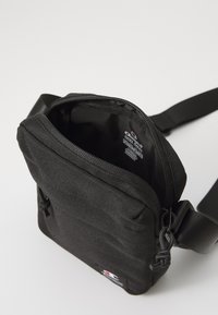 Sac à épaule en tissu noir avec fermeture éclair, sangle réglable et badge logo extérieur. Doublure intérieure lisse, sans poches supplémentaires.