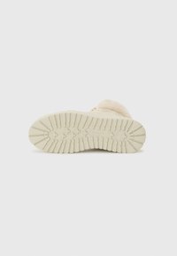 Semelle d'une botte beige clair avec un fond en caoutchouc texturé, présentant des nervures et des motifs pour une adhérence, sur un fond blanc uni.
