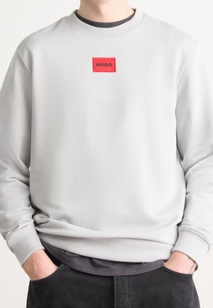 Lichtgrijze sweatshirt met een rode rechthoekige patch met het label "HUGO" gedragen over een zwart shirt, met handen in de zakken van donkere broeken.