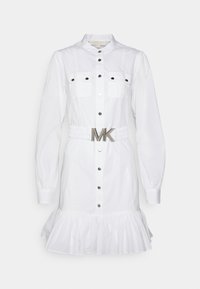 Robe blanche à manches longues boutonnée avec poches poitrine, ourlet à volants et une large ceinture avec une grande boucle métallique MK.