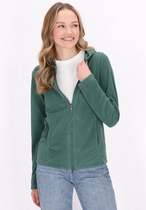Felpa verde con zip, dotata di cappuccio attaccato e maniche lunghe, abbinata a una maglietta bianca e jeans in denim.