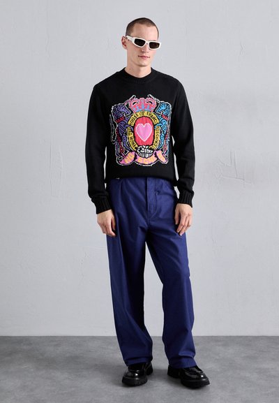 Pull en tricot noir avec un design graphique coloré et du texte, associé à un pantalon ample bleu marine et des chaussures à plateformes noires. Lunettes de soleil blanches portées.