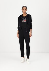 Sudadera negra con acentos metálicos, camiseta gráfica negra y pantalones deportivos negros con detalles laterales. Zapatillas blancas completan el conjunto.