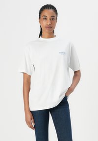 Balta medvilnė t-shirt su laisvu siluetu, ant kairiojo krūtinės puikuojasi mažas mėlynas "GUESS" logotipas. Suderinta su tamsiais džinsais.