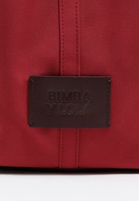 Czerwony plecak z tkaniny, z prostokątną, brązową skórzaną metką wytłoczoną z napisem "BIMBA Y LOLA." Widoczne detale przeszycia podkreślają design.