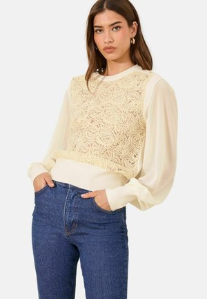 Vrouw met lang bruin haar die gouden oorringen draagt, een doorzichtige crèmekleurige blouse met pofmouwen, een gestructureerde crèmekleurige mouwloze trui en blauwe spijkerbroek.